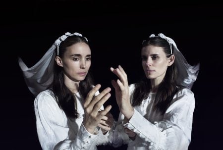 A woman in a white dress and veil touches her own reflection in a mirror against a dark background.