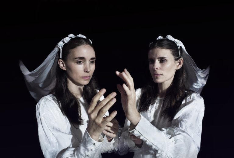 A woman in a white dress and veil touches her own reflection in a mirror against a dark background.