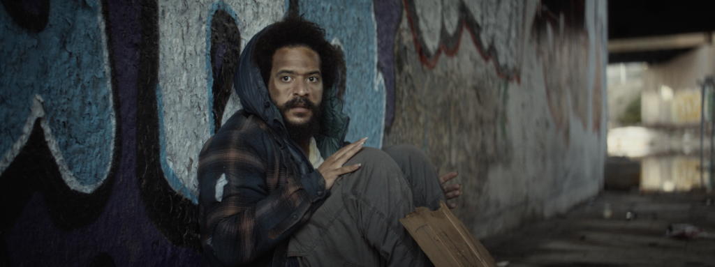 A man with curly hair and a beard sits against a graffiti-covered wall under a bridge, holding a paper bag and looking toward the camera.