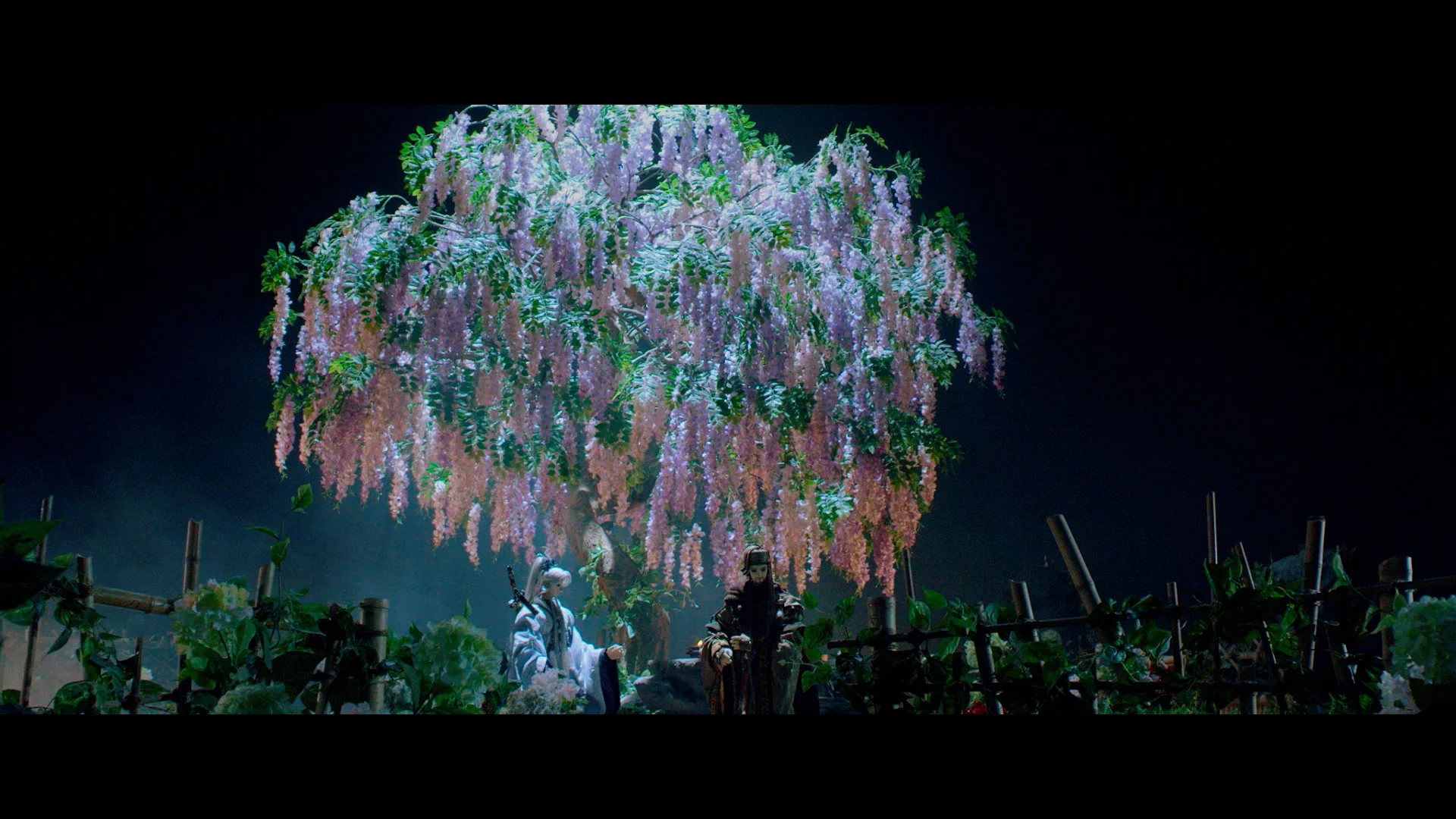 Two people in traditional clothing stand under a large, blooming tree with cascading purple flowers, surrounded by greenery at night.
