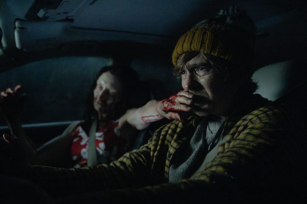 A man wearing a yellow knit hat drives a car at night, looking worried, while a woman with blood on her arm sits in the passenger seat.
