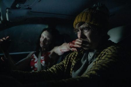 5 Confirmed Resident Evil Movie Easter Eggs You Might Have Missed A man wearing a yellow knit hat drives a car at night, looking worried, while a woman with blood on her arm sits in the passenger seat.