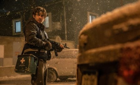 A person holding a rifle and a medical bag stands outdoors in falling snow at night, looking toward a vehicle in the foreground.