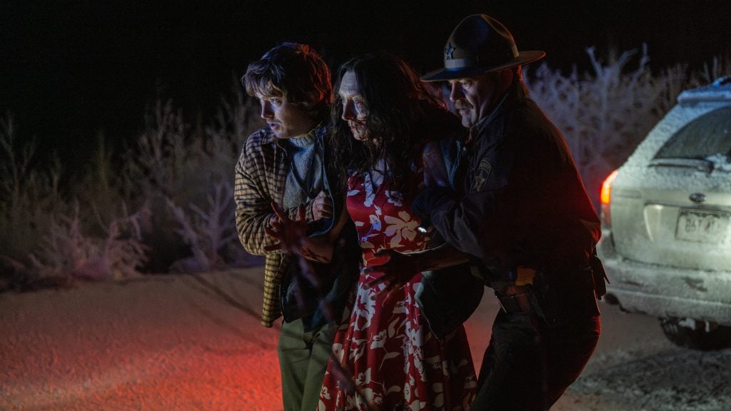 A bloodied woman in a red dress is helped by a man and a police officer near a frosty road at night beside a car with its lights on.