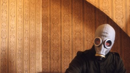 Person wearing a gas mask and dark clothing sits indoors against patterned brown wallpaper under an arched ceiling.