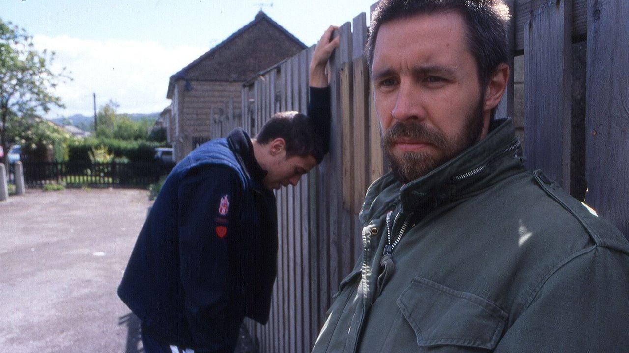 Two men stand outside by a wooden fence; one faces the camera with a serious expression, while the other leans with his head down against the fence in the background.