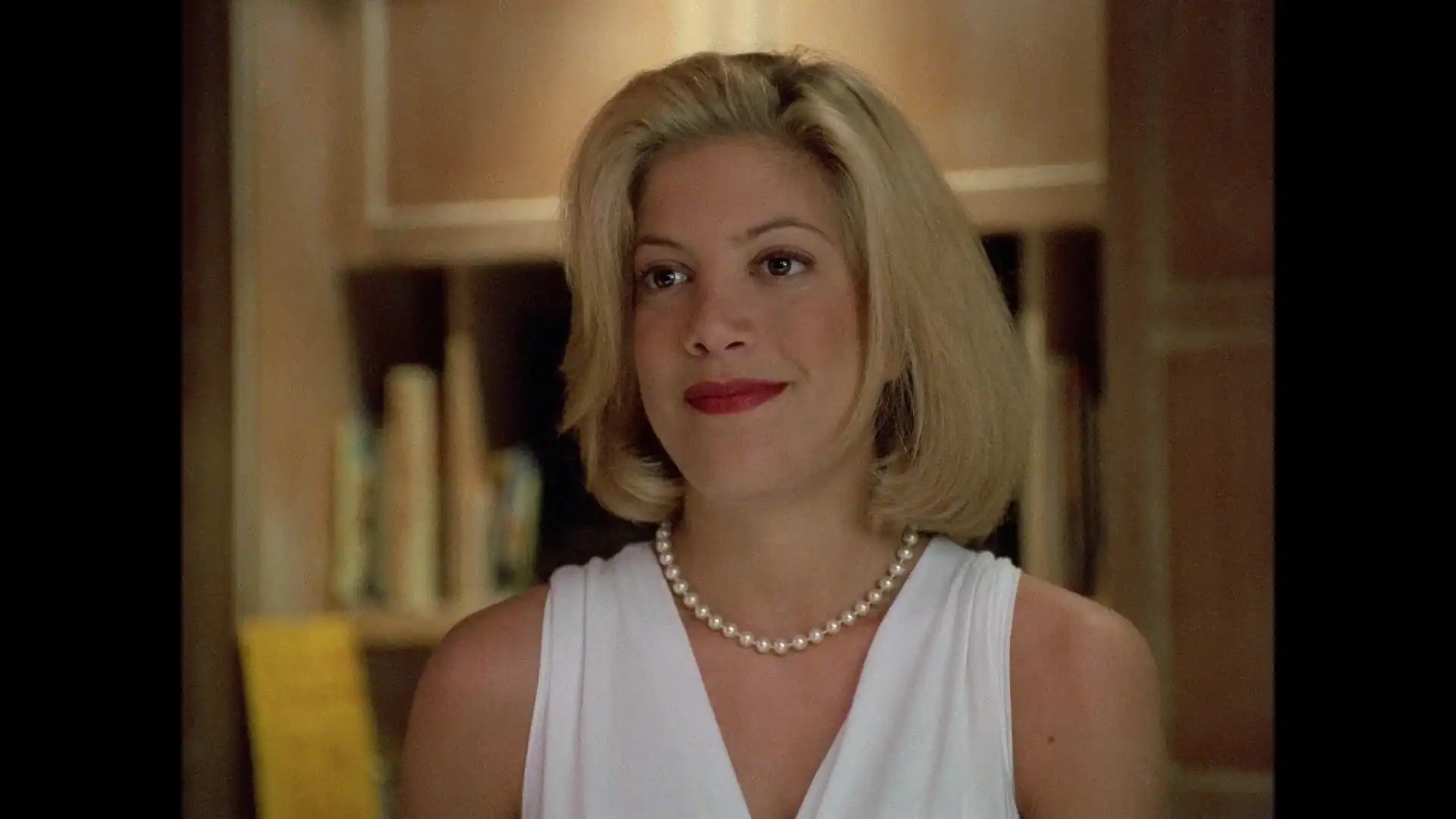 Woman with short blonde hair wearing a white sleeveless top and pearl necklace stands indoors in front of shelves with books.