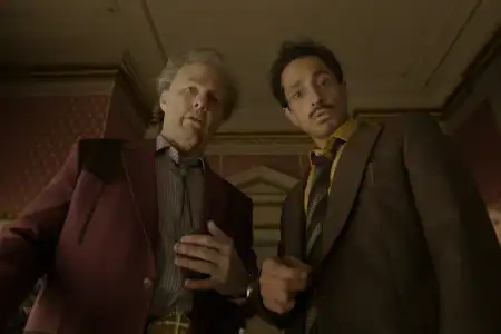 Two men in suits look downward, standing in a richly decorated room with patterned wallpaper and ornate molding, as seen from a low-angle perspective.