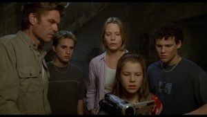 Five people stand close together, looking with concern at a video camera being held and operated by a young girl in the foreground.