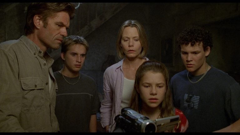 Five people stand close together, looking with concern at a video camera being held and operated by a young girl in the foreground.