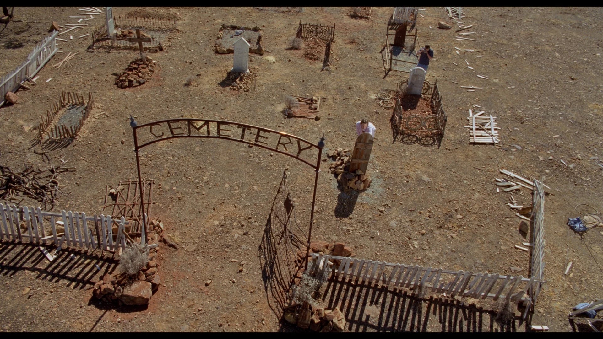 Aerial view of a sparse, old cemetery with simple graves, uneven fencing, rocky ground, and three people standing among the headstones.