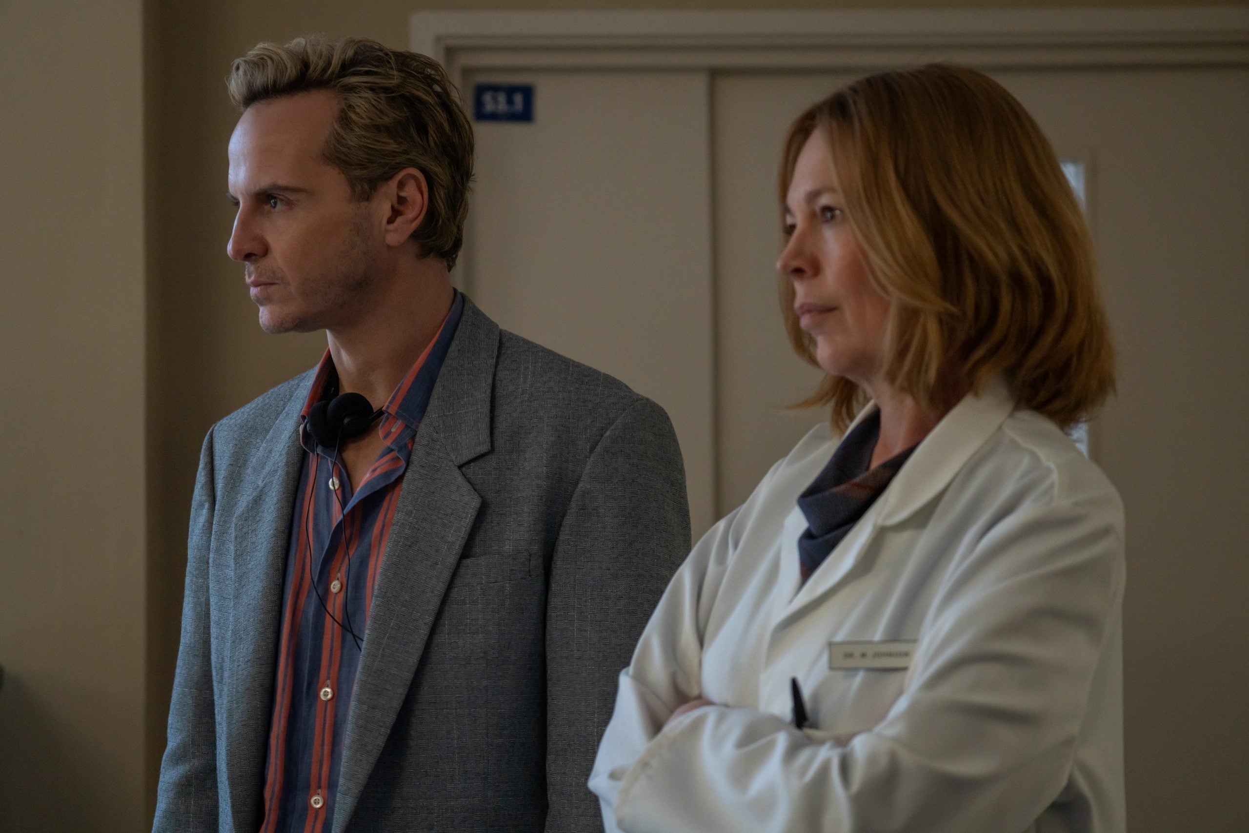 A man in a blazer and a woman in a white lab coat stand side by side indoors, looking intently in the same direction.