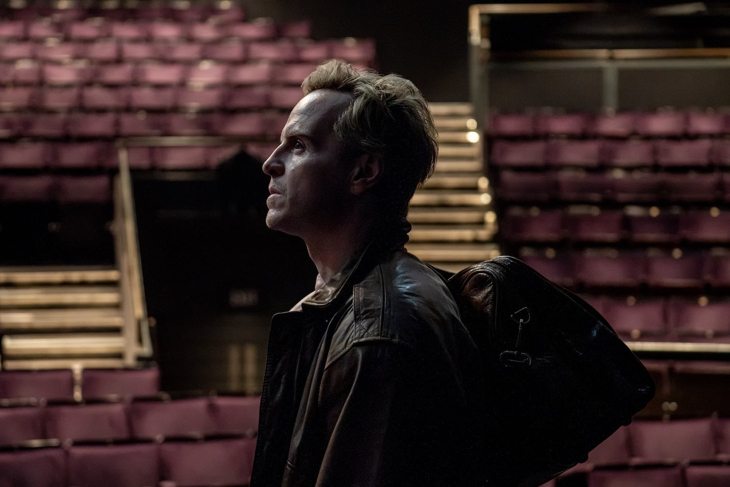 A man with a bag over his shoulder stands in profile in an empty theater with rows of maroon seats behind him.