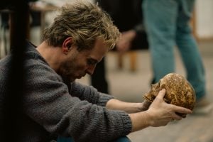 A man kneels indoors, holding and looking intently at a human skull. Other people are visible in the blurred background.