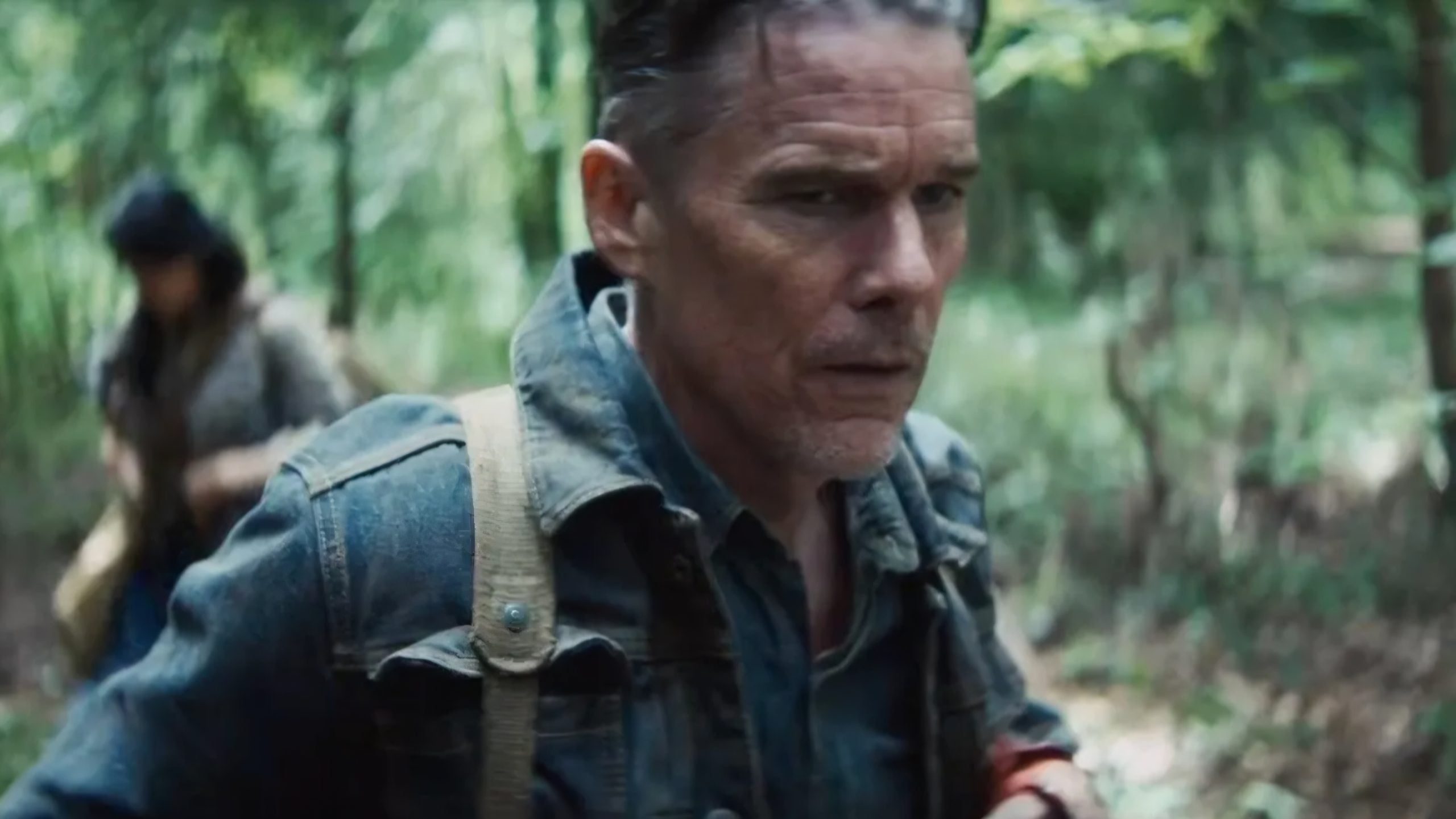 A man in a denim jacket walks through a wooded area, with another person slightly out of focus behind him.