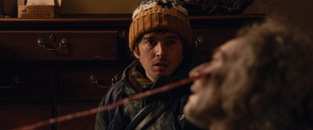 A person wearing a knit hat looks concerned while another person in the foreground has a long object protruding from their mouth.