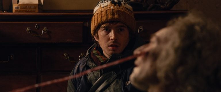 A person wearing a knit hat looks concerned while another person in the foreground has a long object protruding from their mouth.