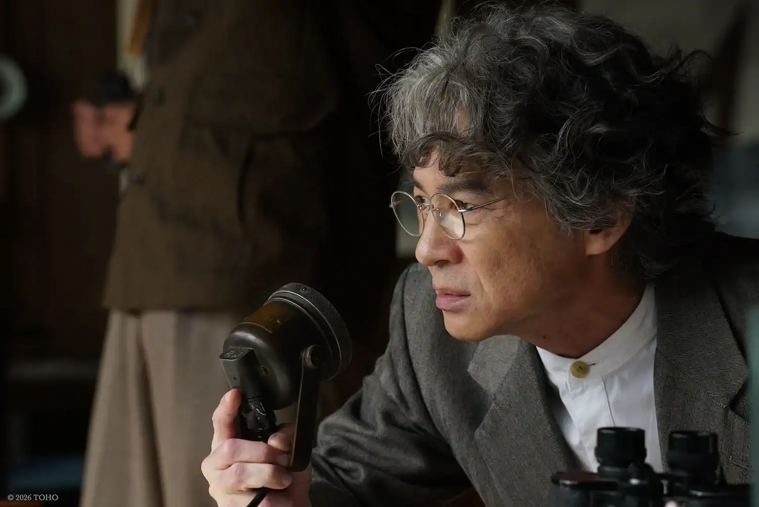 An older man with gray curly hair and glasses holds a vintage microphone, wearing a suit and white shirt, with binoculars on the table in front of him. Another person is blurred in the background.
