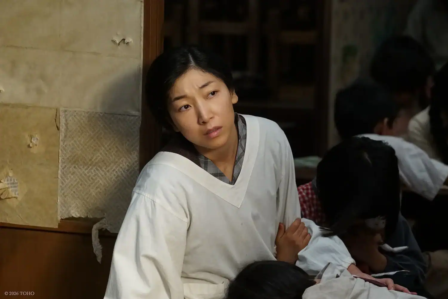 A woman in a white robe sits on the floor, leaning against a wall, looking pensive. Several children rest against her, partially out of frame.
