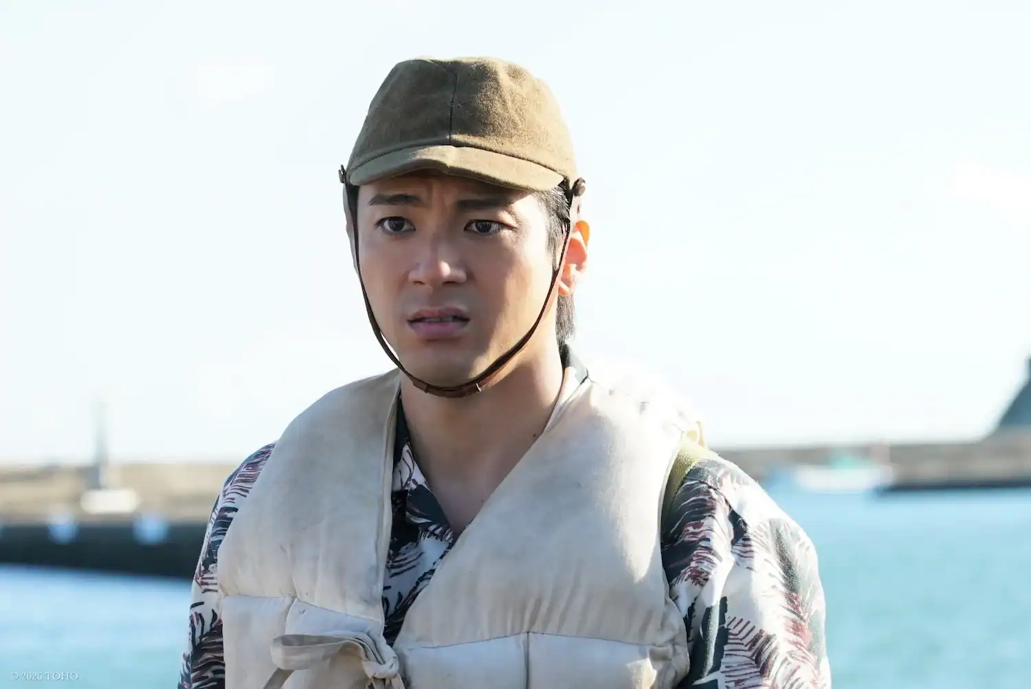 A man wearing a brown cap, floral shirt, and life vest stands outdoors near water, looking concerned.