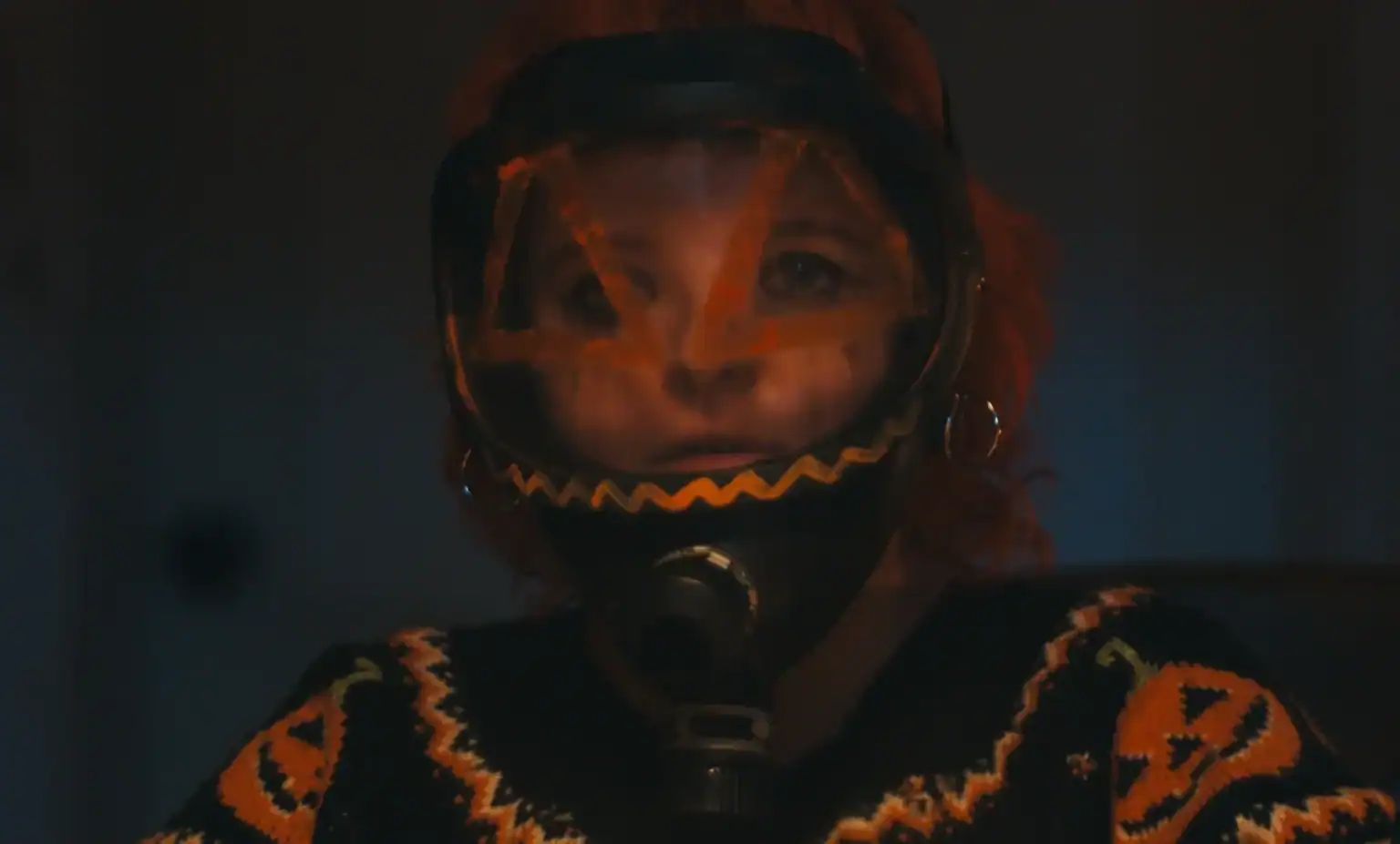 ‘Hallowarrior’ Review – A Haunted ‘Home Alone’ That Is Autumnal Eye-Candy Person with orange hair wears a gas mask decorated with a jack-o’-lantern face and a black and orange Halloween-themed sweater.