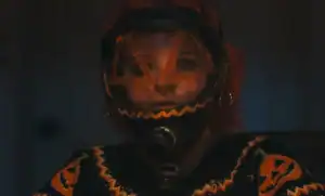 Person with orange hair wears a gas mask decorated with a jack-o’-lantern face and a black and orange Halloween-themed sweater.