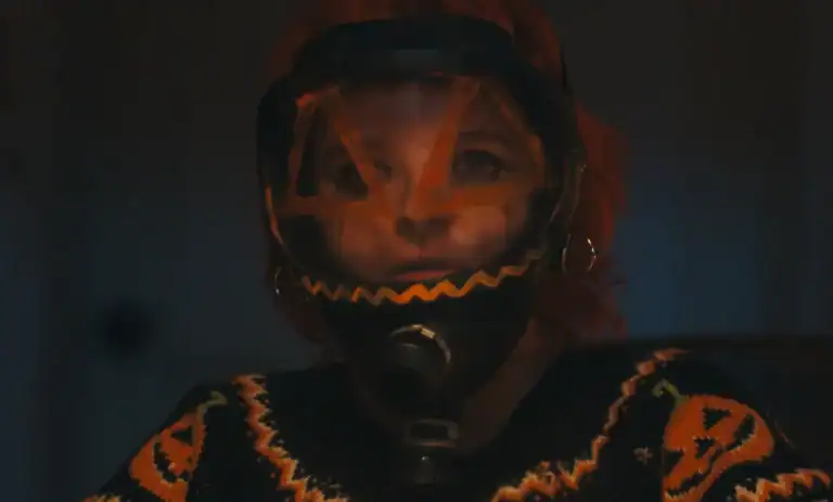 Person with orange hair wears a gas mask decorated with a jack-o’-lantern face and a black and orange Halloween-themed sweater.