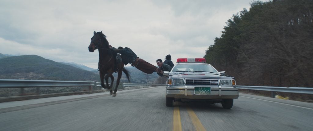 A person on horseback leaps next to a moving police car on a mountain road, with trees on the right and hills in the background.
