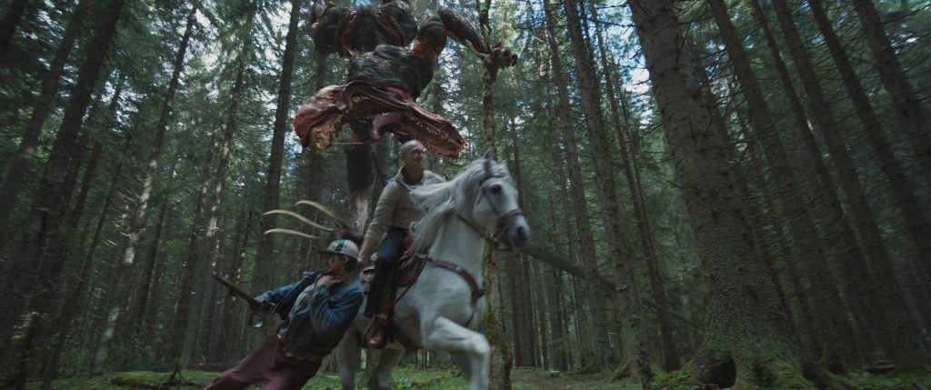 A monstrous creature leaps behind a man on horseback and another man with a rifle in a dense forest during daylight.