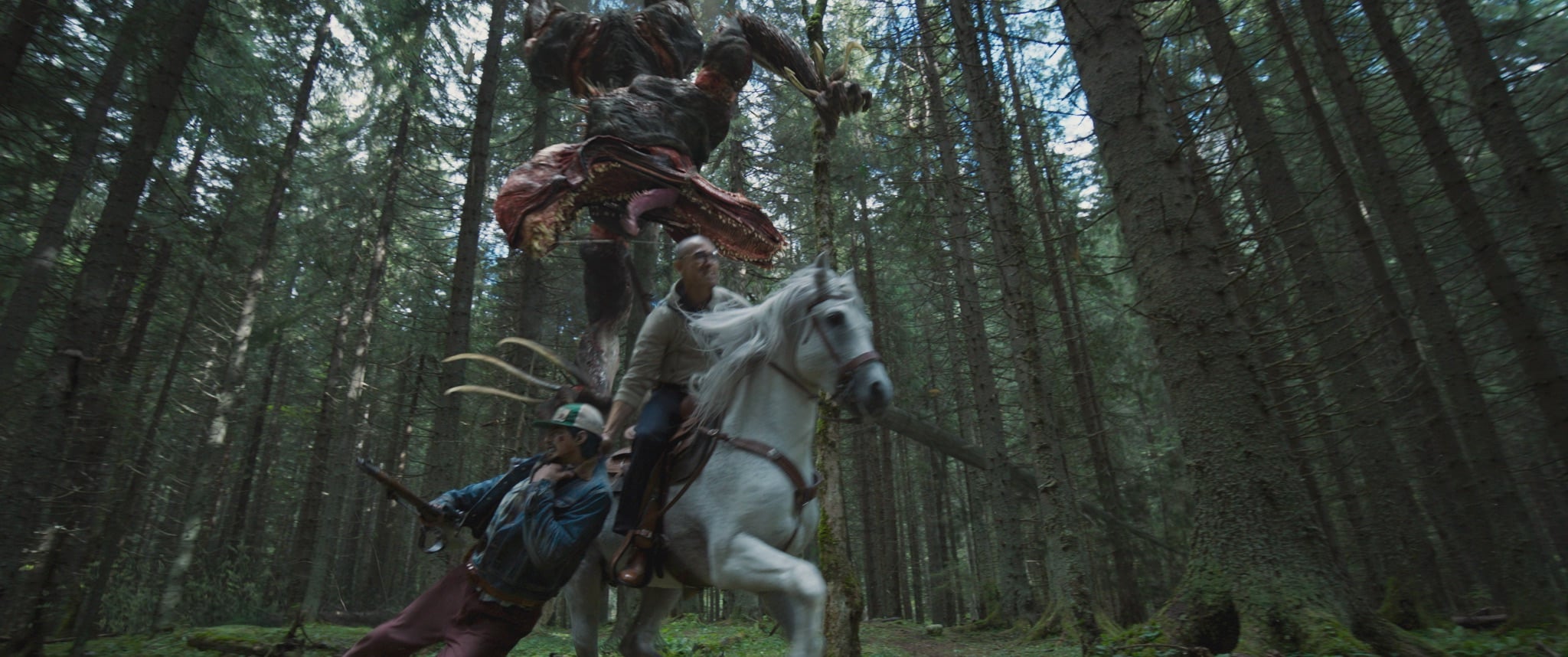 A monstrous creature leaps behind a man on horseback and another man with a rifle in a dense forest during daylight.