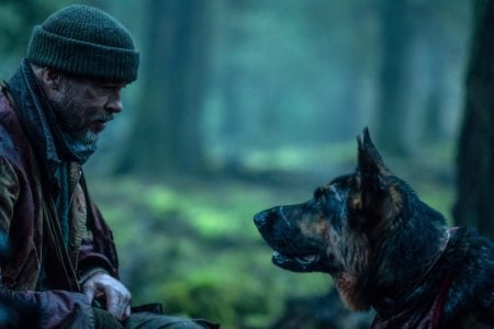 ‘Heart of the Beast’ Review – A Harrowing Tale Of Man And Man’s Best Friend Battling The Brutality Of Nature A man in a beanie and rugged clothing sits in a forest facing a German Shepherd dog. The background is dense with trees and greenery.