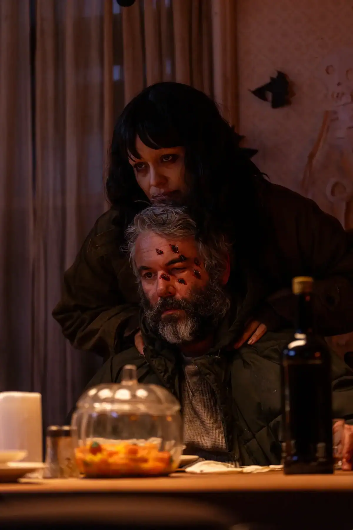 A woman stands behind a seated man with wounds on his face at a dimly lit table set with a bottle, cups, and a bowl of food.