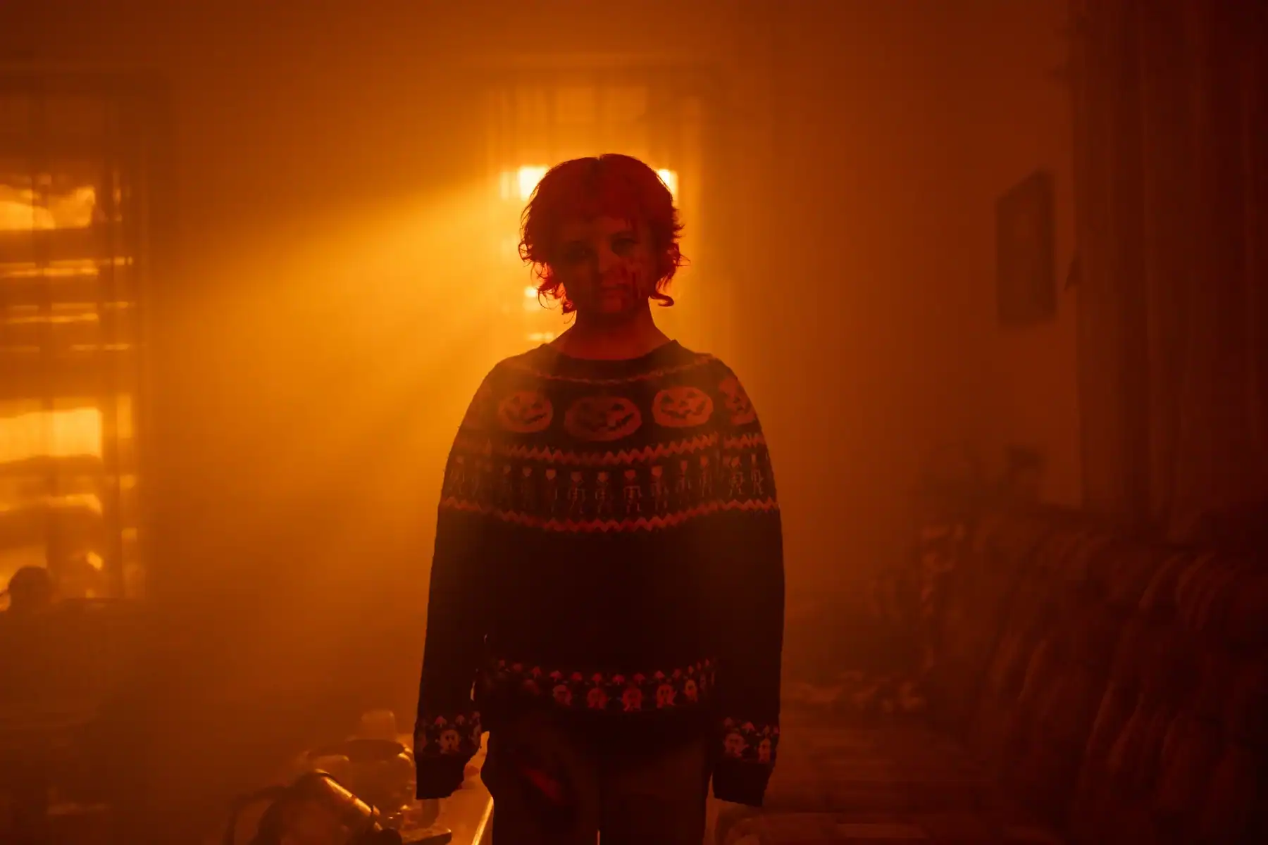 A person with a bloodied face and red hair stands in a dimly lit room, wearing a sweater with jack-o’-lantern designs. Sunlight streams through windows behind them.