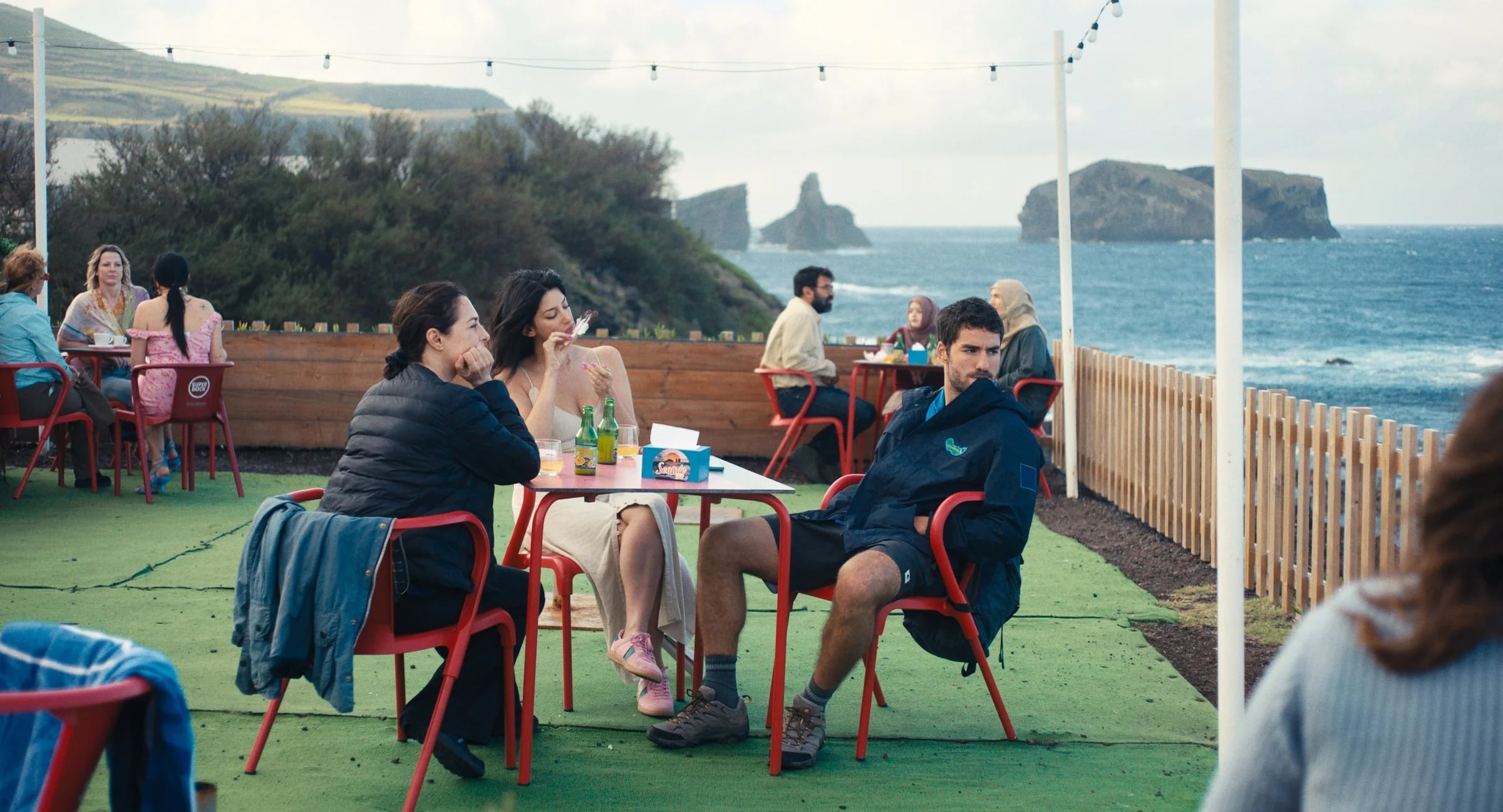 People sit at red tables and chairs on an outdoor patio near the ocean, with cliffs and islands in the background. Some are eating and drinking, and string lights are overhead.