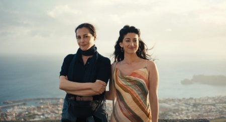 ‘Honeyjoon’ Blu-Ray Review – An Emotional, Darkly Comedic Gem That Finds Levity In Grieving Two women stand side by side outdoors with a scenic view of a coastal town and water in the background. One woman wears a dark outfit and scarf; the other wears a striped dress.