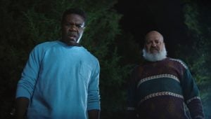 Two men stand outside at night, looking downward with concerned expressions, against a backdrop of trees and darkness.