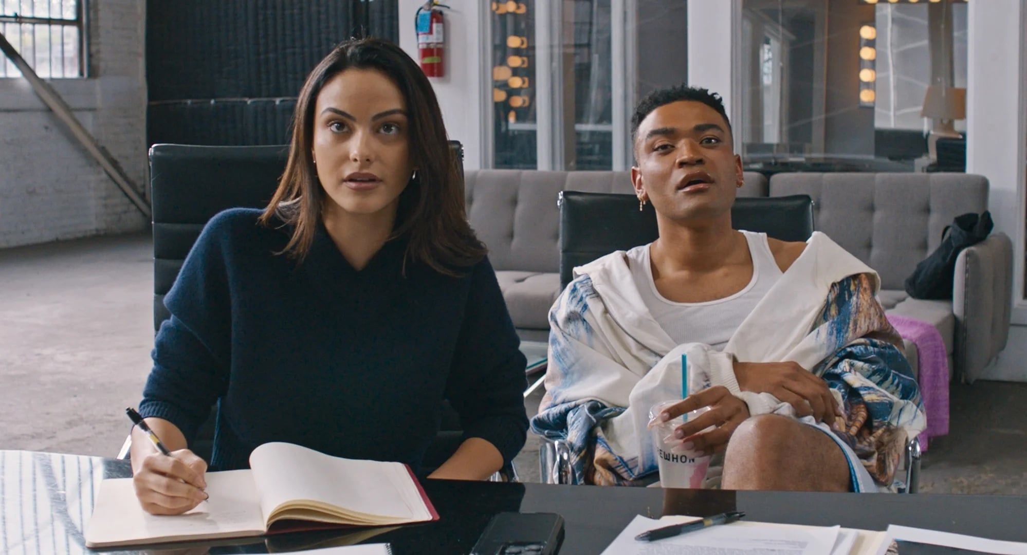 Two people sit at a table with papers and notebooks, looking forward with focused expressions. One holds a drink while the other has an open notebook.