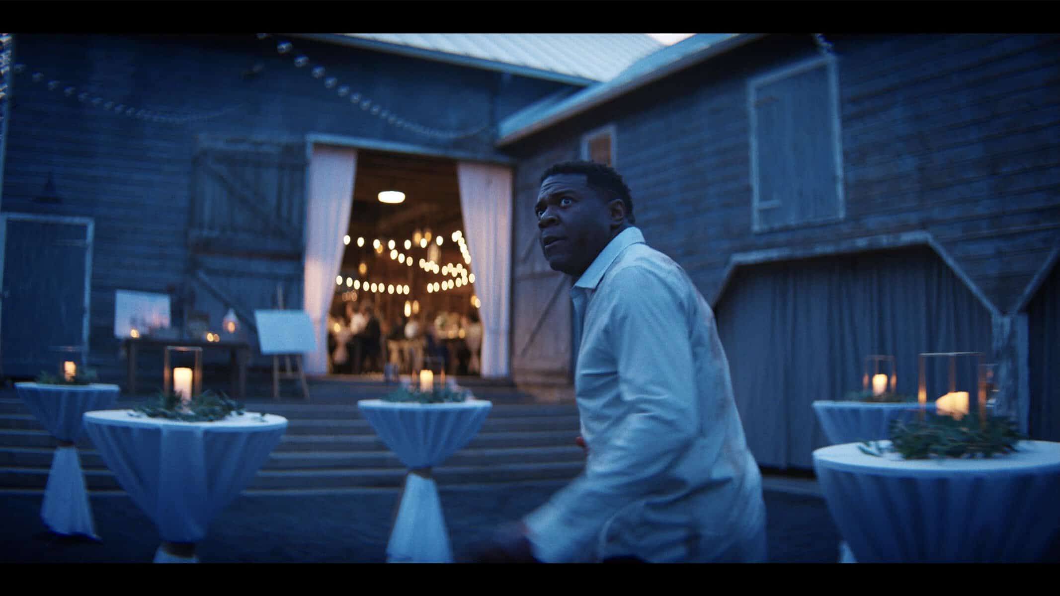 A man in a white shirt stands outside a barn decorated with string lights and cocktail tables, looking up with a concerned expression.