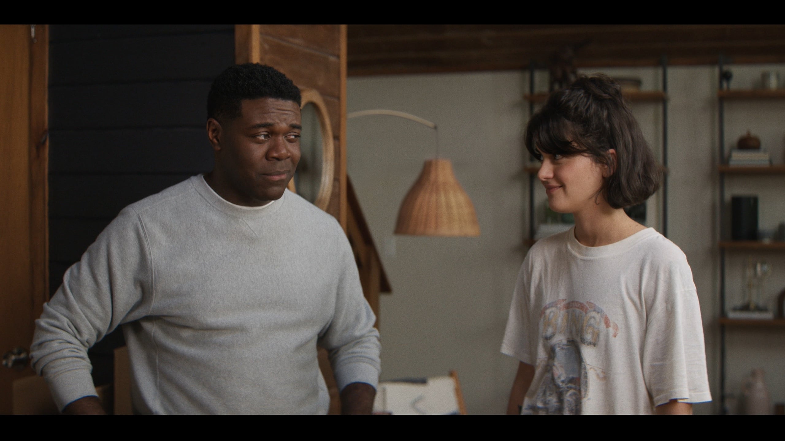 A man in a gray sweater and a woman in a white T-shirt stand indoors, facing each other and smiling slightly, with shelves and decor visible in the background.