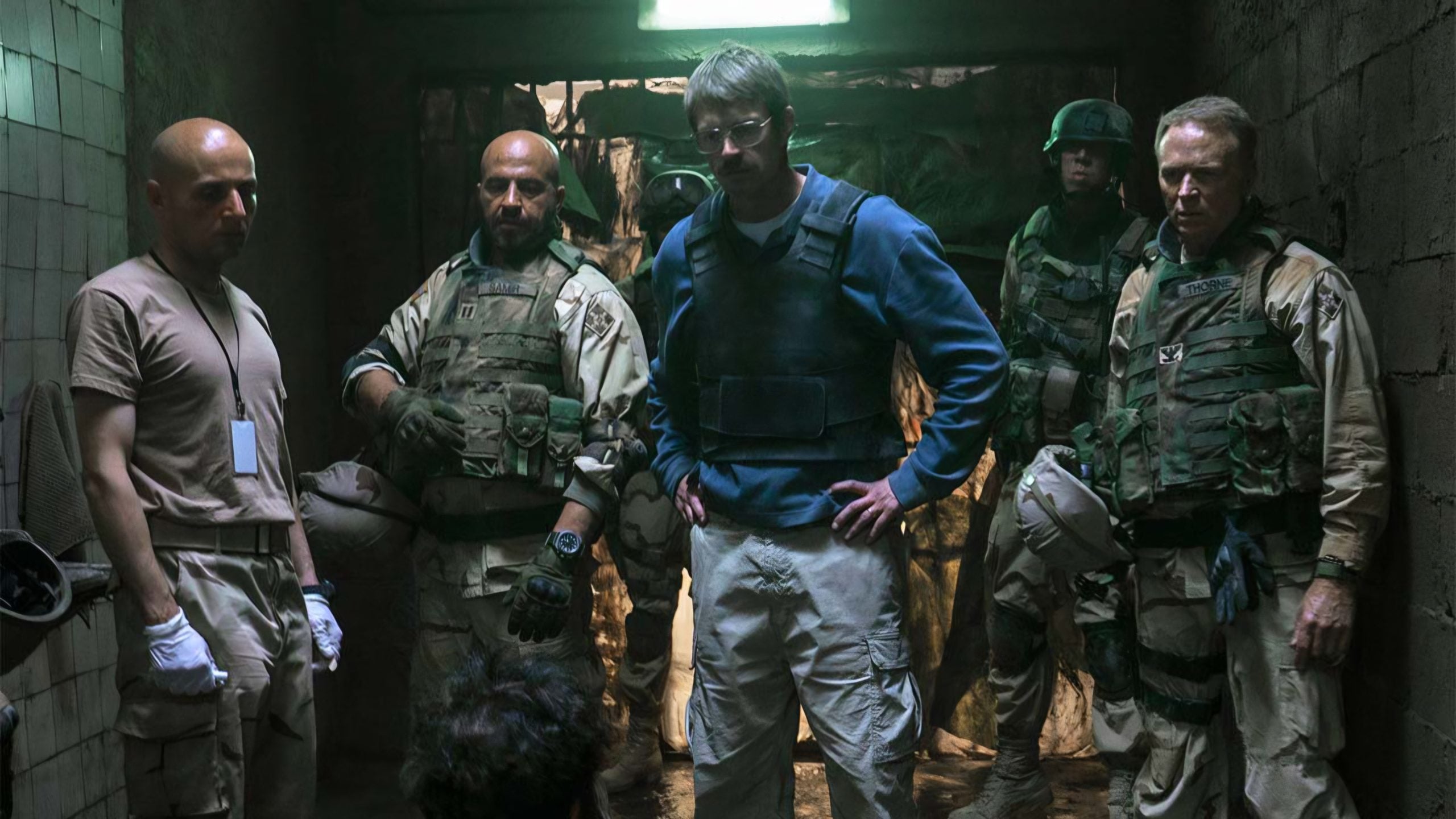 Five men in military gear stand in a dimly lit, worn-out corridor, facing a person kneeling on the ground. A bright light shines overhead, casting shadows on the walls.