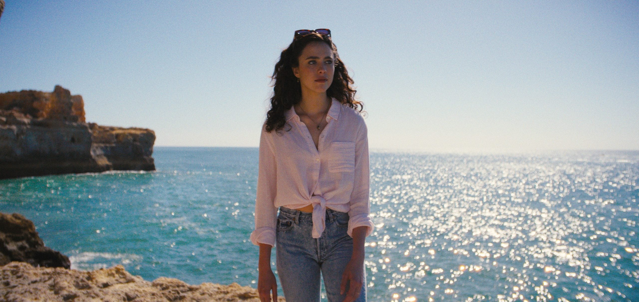 A woman in a light shirt and jeans stands on a rocky shoreline with the ocean and cliffs in the background under a clear sky.