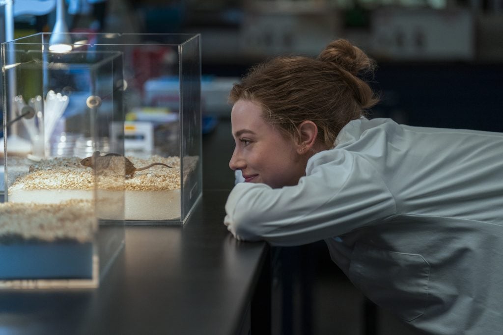 A woman in a white lab coat smiles while looking at a mouse inside a glass enclosure on a lab table.