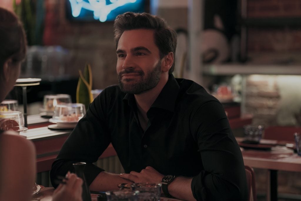 Man with a beard and dark shirt sits at a restaurant table, facing another person, with a slight smile in a dimly lit setting.