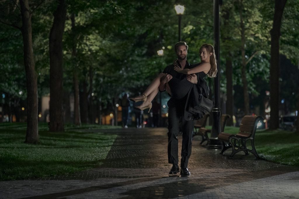 A man in a suit carries a woman in a black dress through a park at night; empty benches and trees line the walkway.