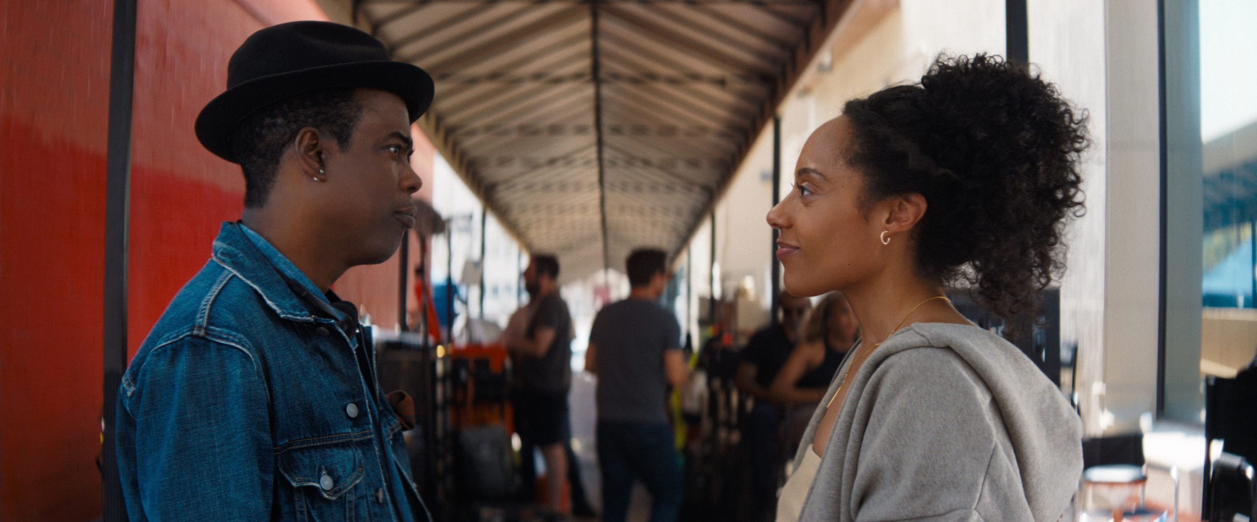 A man in a denim jacket and black hat faces a woman in a hoodie. They stand outdoors under a covered walkway, with people and tables in the background.