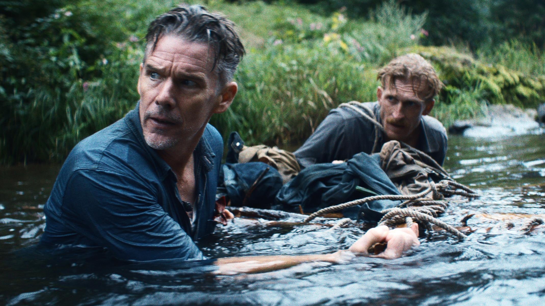 Two men wade through a shallow river, clinging to their gear on a makeshift raft, surrounded by greenery.