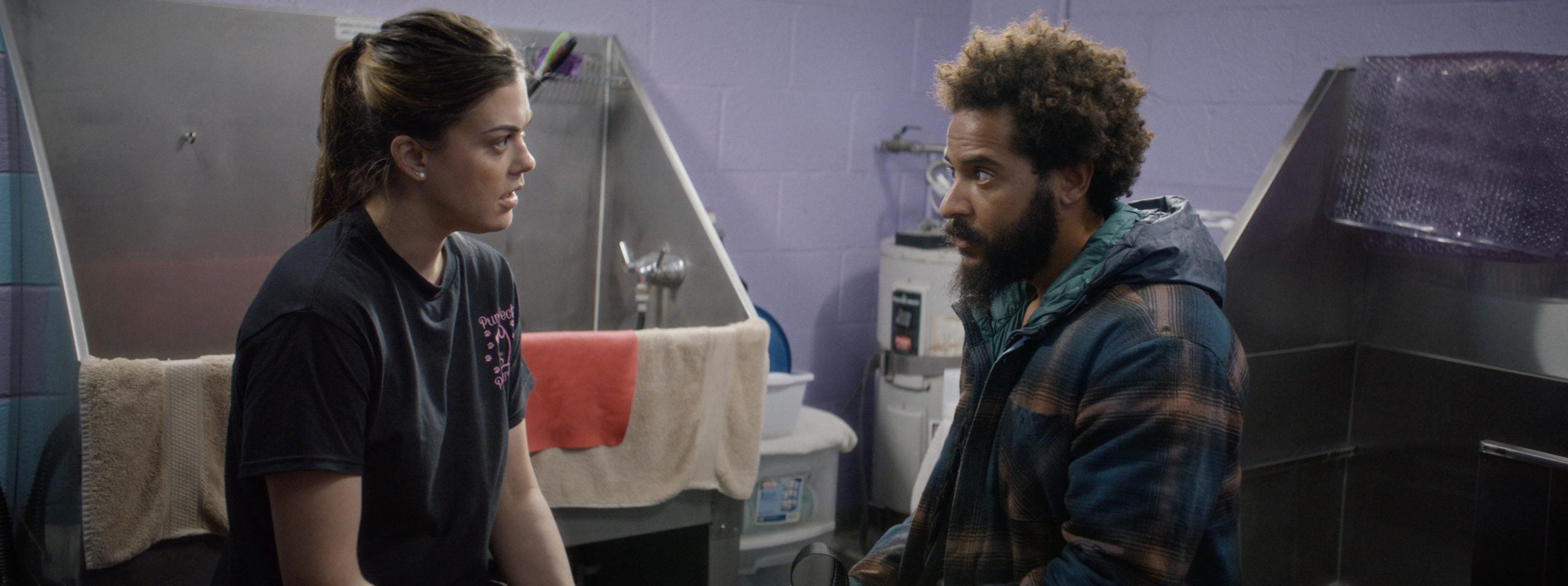 A woman and a man sit facing each other in a room with lavender walls and metal sinks, appearing to have a serious conversation.