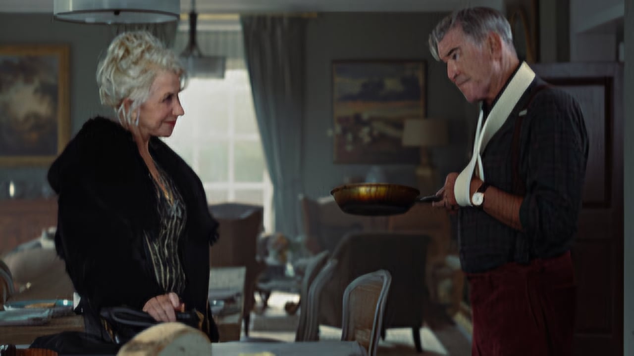 An older woman in a dark outfit faces an older man with a sling, holding a frying pan in a cozy, well-furnished room with paintings and a dining table.