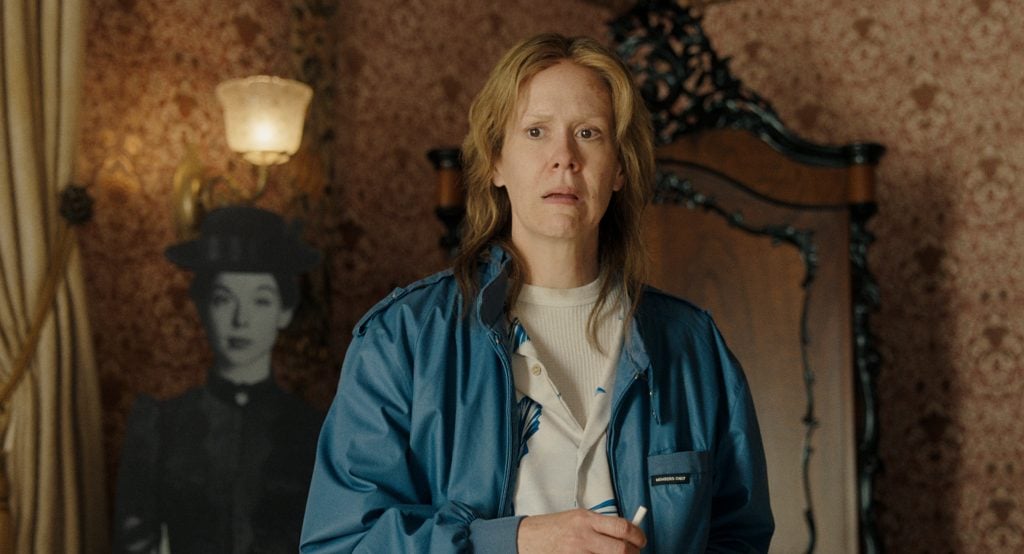 A person in a blue jacket stands indoors, holding a cigarette, with a cardboard cutout of a woman in the background against patterned wallpaper.