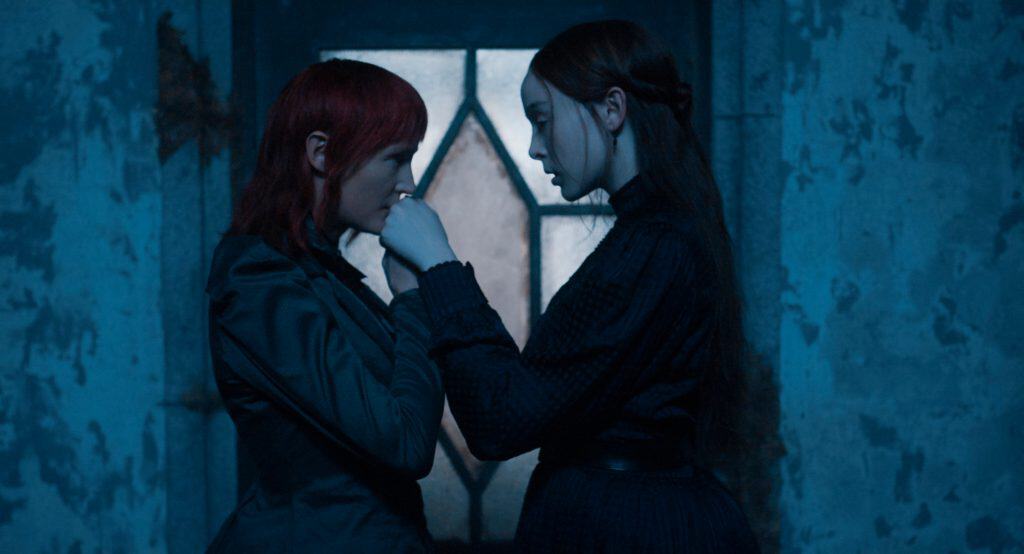 Two women in dark clothing stand close together indoors, one gently holding the other’s hand to her lips, with a blue-lit window and textured wall in the background.